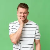 Mężczyzna z bólem zęba ściska policzek, szukając ulgi, jakby używał zamrażacza na ból zęba.