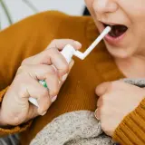 Kobieta aplikuje spray do gardła, łagodząc ból związany z aftami na migdałach. Zdjęcia pokazują jej dłoń z aplikatorem.