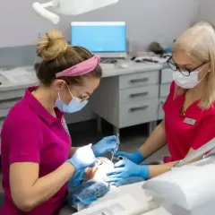 Dwie dentystki w maseczkach i rękawiczkach wykonują zabieg. Jedna z nich stosuje znieczulenie nasiękowe, by zapewnić pacjentowi komfort.