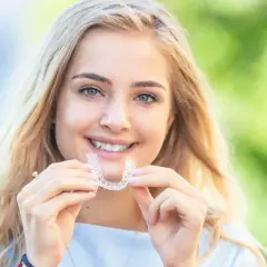 Uśmiechnięta blondynka trzyma w dłoniach przezroczysty aparat retencyjny, symbol pięknego uśmiechu.