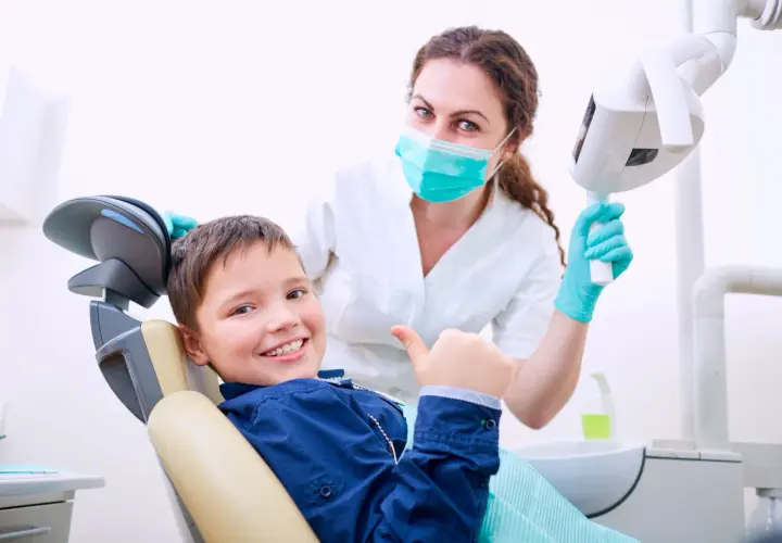 Uśmiechnięty chłopiec daje kciuk w górę w gabinecie stomatologicznym. Uśmiechnięta dentystka w masce i rękawiczkach stoi za nim.