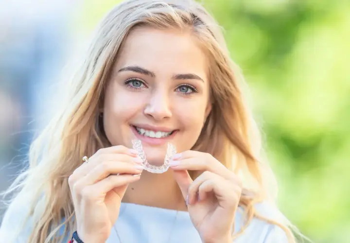 Uśmiechnięta blondynka trzyma w dłoniach przezroczysty aparat retencyjny, symbol pięknego uśmiechu.