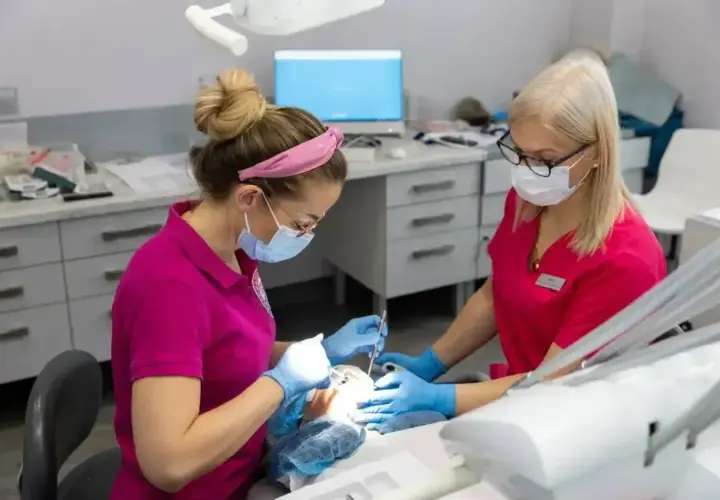 Dwie dentystki w maseczkach pracują przy pacjencie. Jedna z nich podaje narzędzia, druga wykonuje zabieg, stosując lidokainę znieczulenie miejscowe.