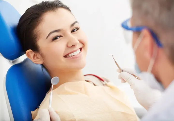 Uśmiechnięta pacjentka na fotelu dentystycznym, podczas gdy dentysta przygotowuje się do zabiegu z użyciem materiału glasjonomer.