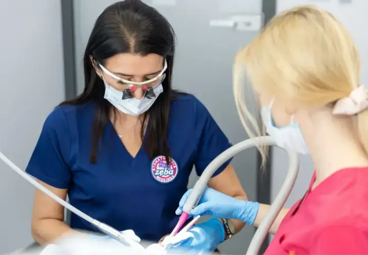 Dentystka w okularach powiększających i masce wykonuje zabieg. Asystentka trzyma ssak. Znieczulenie śródwięzadłowe zapewnia komfort pacjentowi.