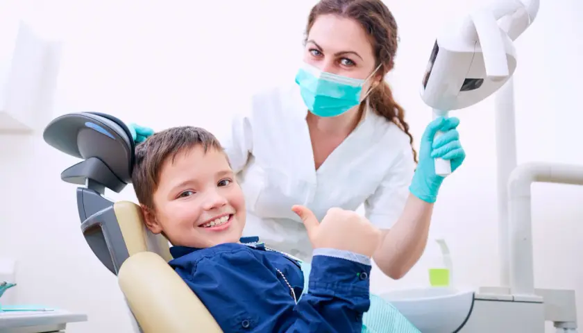 Uśmiechnięty chłopiec daje kciuk w górę w gabinecie stomatologicznym. Uśmiechnięta dentystka w masce i rękawiczkach stoi za nim.