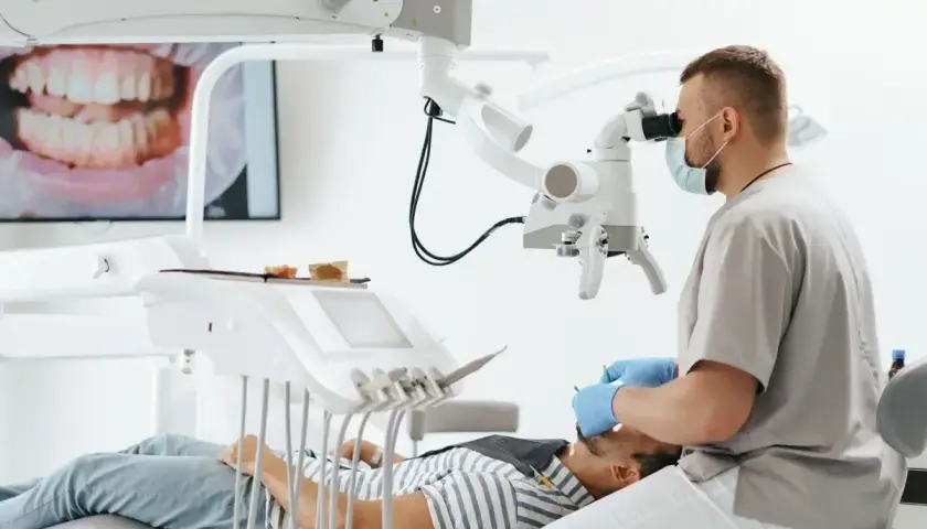 Stomatolog w masce i rękawiczkach bada pacjenta pod mikroskopem. Na ekranie widać zbliżenie zębów.