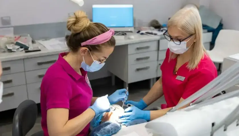 Dwie dentystki w maseczkach pracują przy pacjencie. Jedna z nich podaje narzędzia, druga wykonuje zabieg, stosując lidokainę znieczulenie miejscowe.