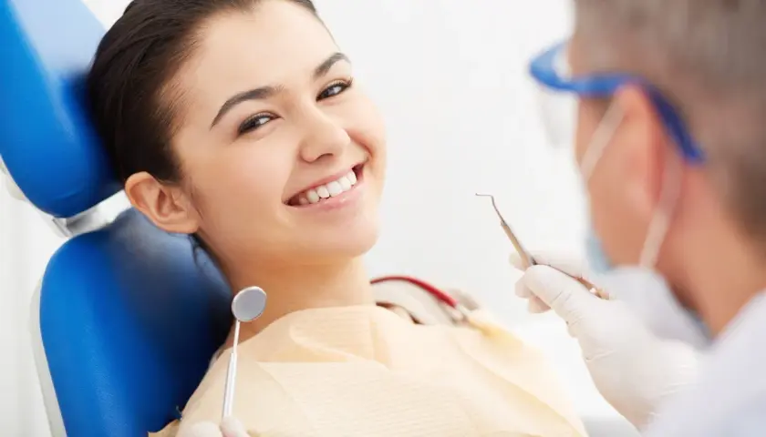 Uśmiechnięta pacjentka na fotelu dentystycznym, podczas gdy dentysta przygotowuje się do zabiegu z użyciem materiału glasjonomer.