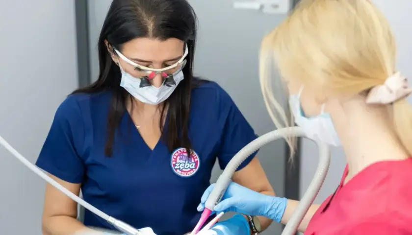 Dentystka w okularach powiększających i masce wykonuje zabieg. Asystentka trzyma ssak. Znieczulenie śródwięzadłowe zapewnia komfort pacjentowi.