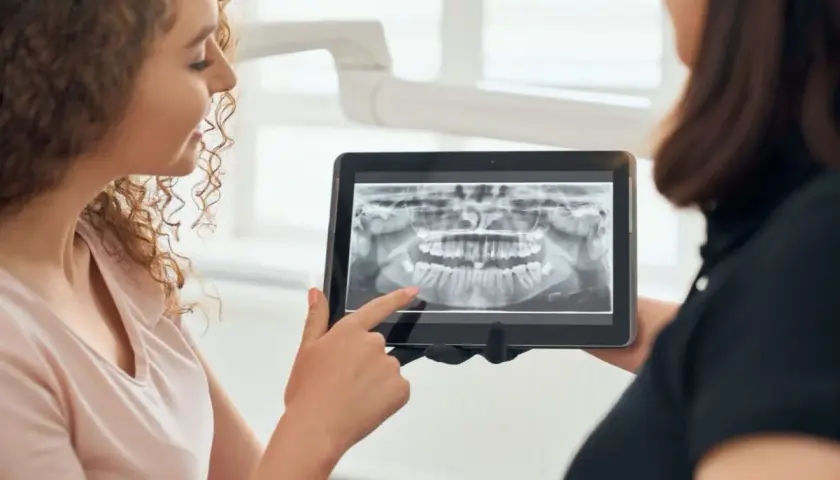 Pacjentka z kręconymi włosami ogląda z dentystką panoramiczny rentgen stomatologiczny na tablecie.
