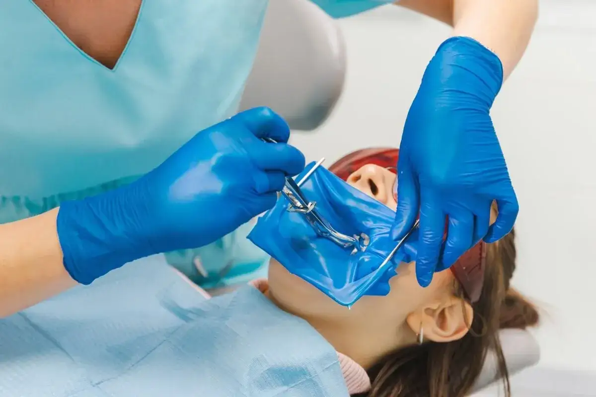 Dentysta zakłada koferdam, izolując pole zabiegowe. Pacjentka leży na fotelu, ma założone okulary ochronne.