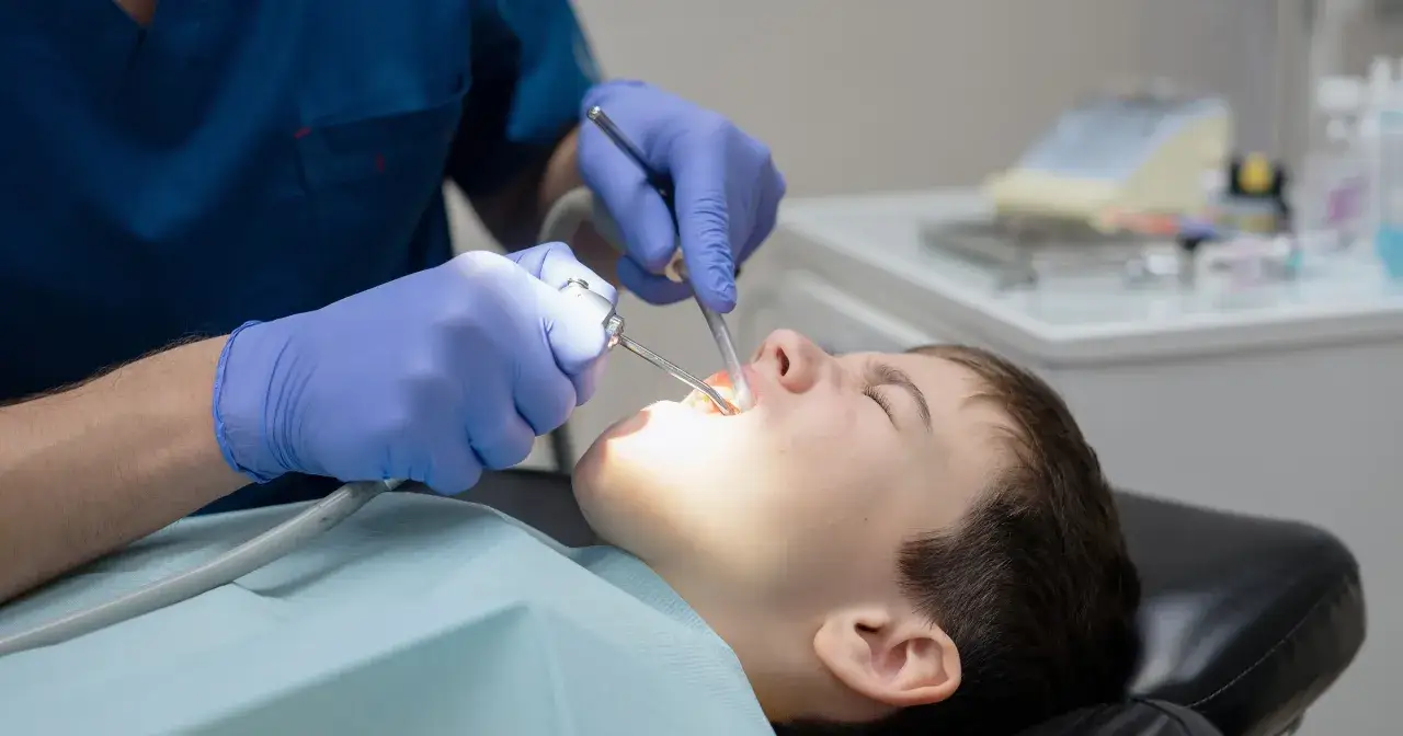 Chłopiec w gabinecie stomatologicznym, poddawany zabiegowi. Dentysta w rękawiczkach używa narzędzi, być może w znieczuleniu ogólnym (narkoza).