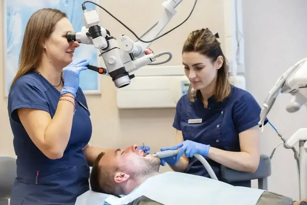 Dentystka z asystentką pracują przy pacjencie. Jedna używa mikroskopu, druga przygotowuje się do zabiegu, który może wymagać narkozy.